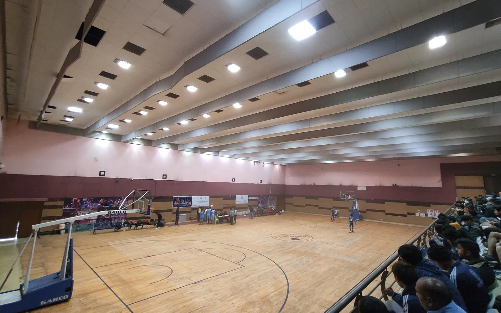 An indoor basketball hall with a wooden court, hoops at both ends and spectators along one side