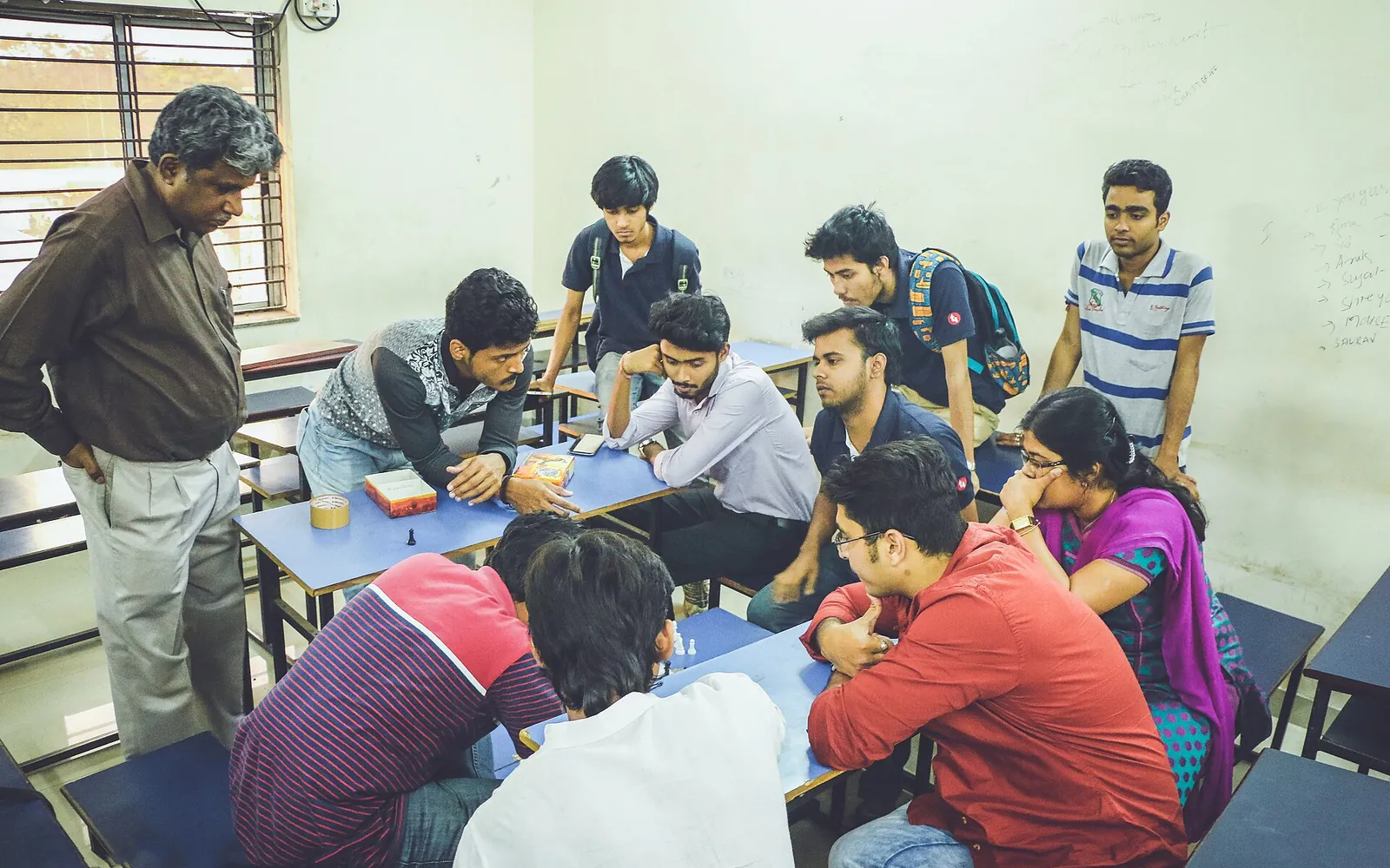 Students gathered around a chess game on a classroom desk, with an older player watching