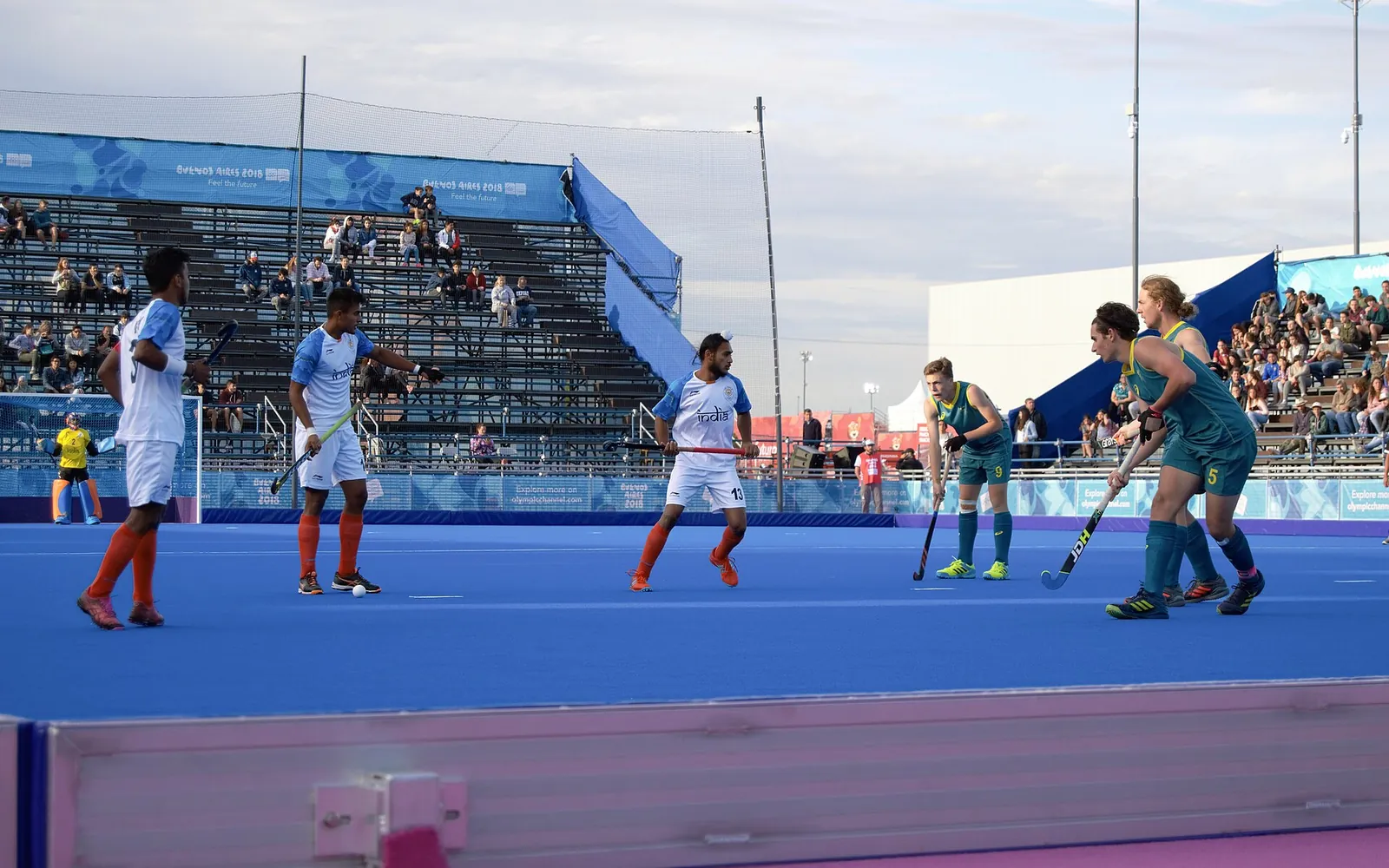 India and Australia junior players with hockey sticks during a match on a blue artificial pitch, with spectators in the stands