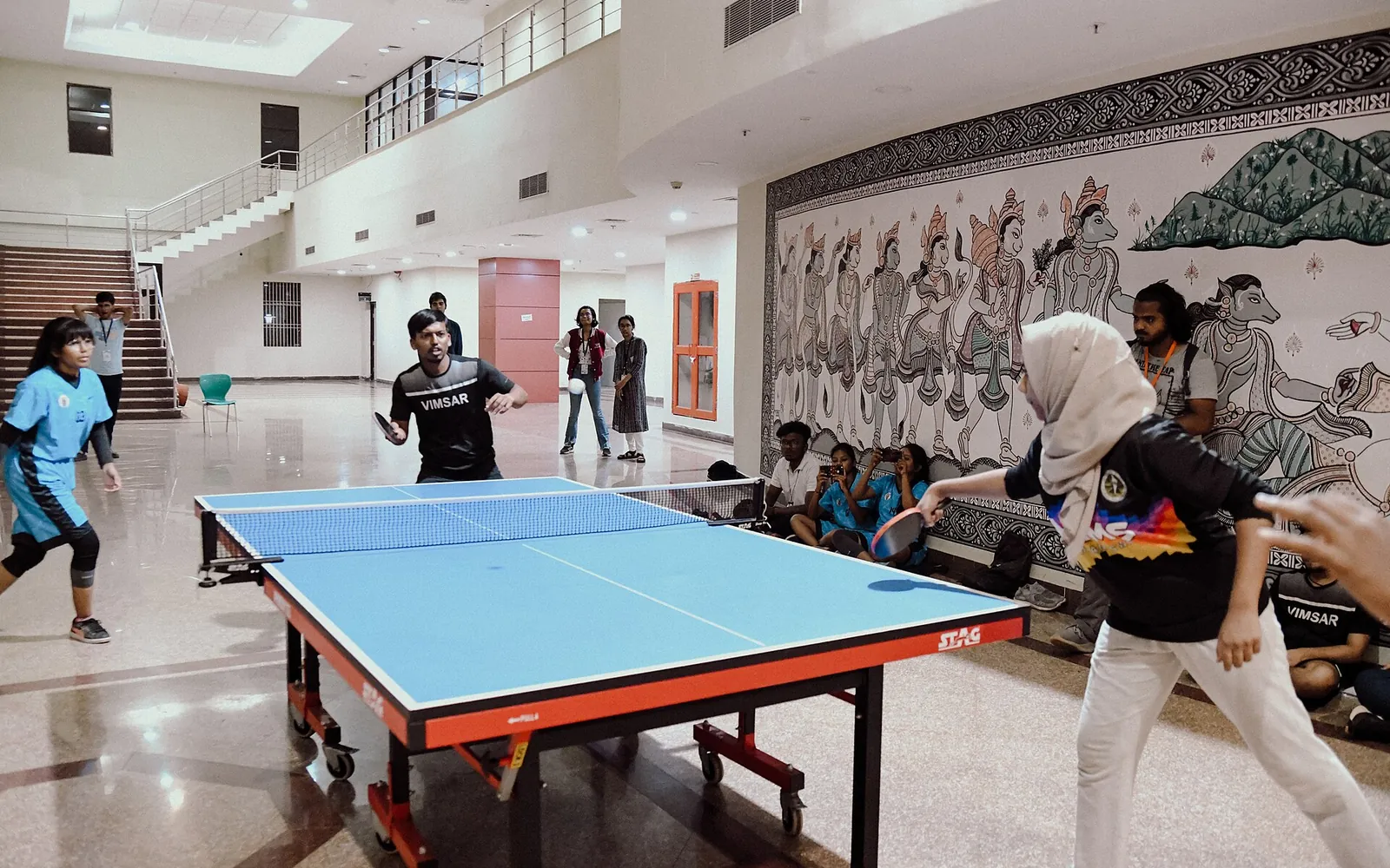 Two players in a rally at a table tennis table inside a large hall