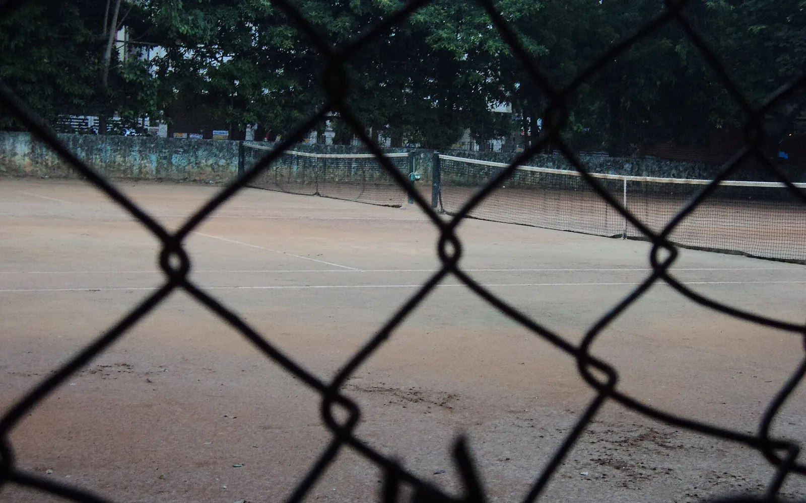 Two empty clay tennis courts with white lines and sagging nets, seen through a chain-link fence